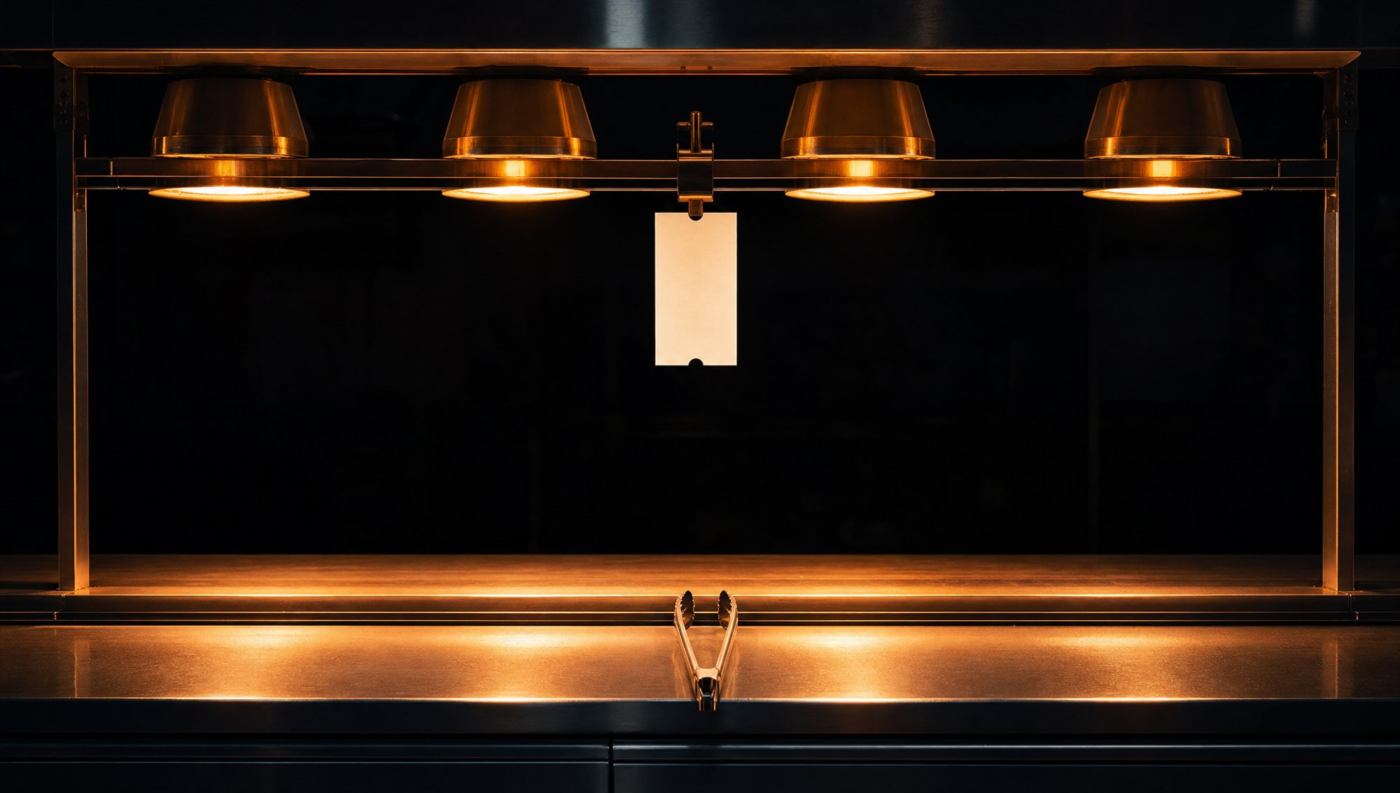 Heat lamps burn over an empty steel pass with one blank ticket clipped dead centre under the light